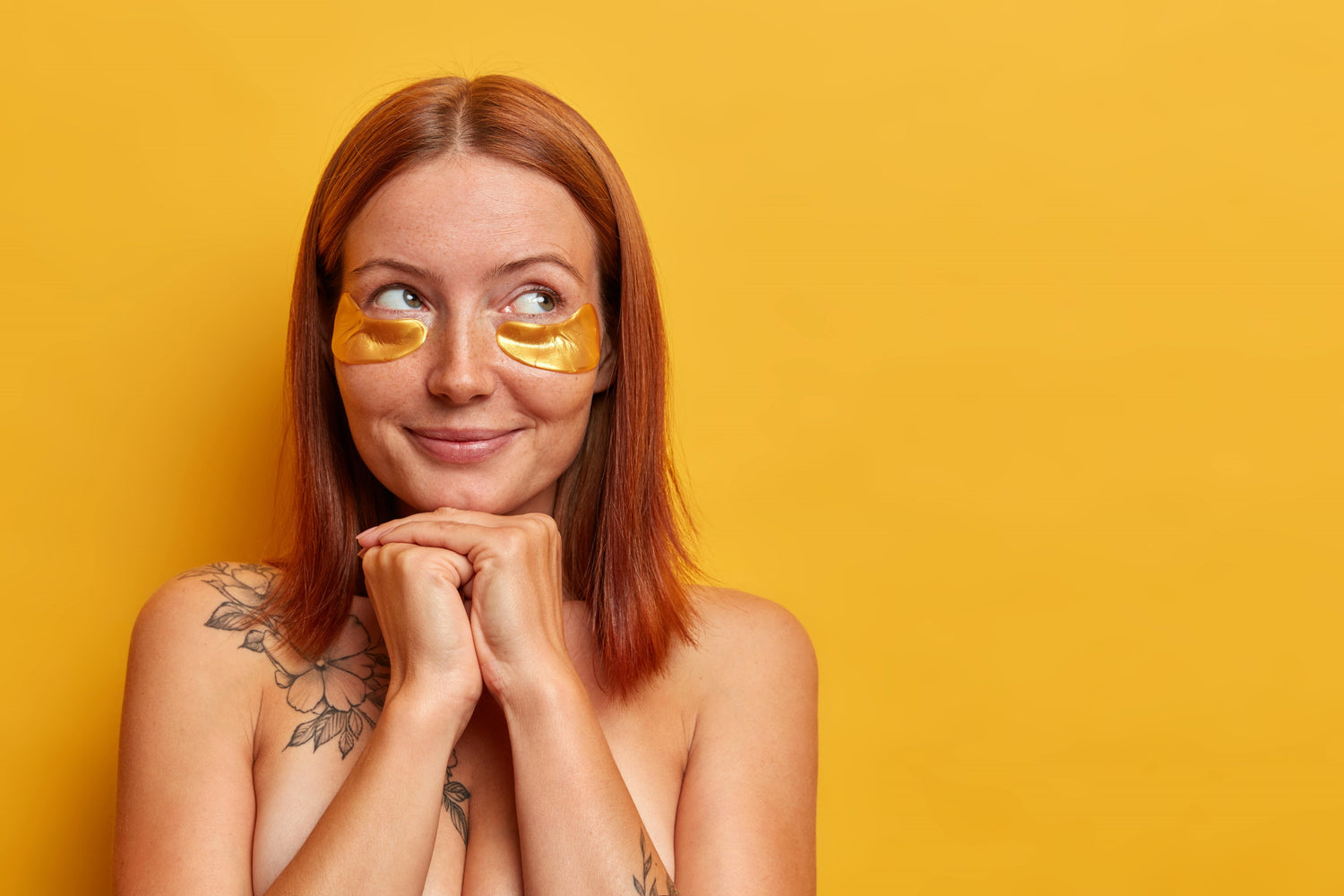 Woman with red hair smiling while wearing gold under-eye patches against a yellow background, featuring article on fine lines and wrinkle prevention