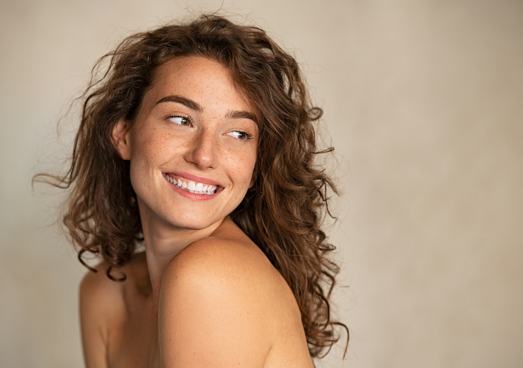 Smiling woman with natural curls looking to the side, showcasing clear, glowing skin, featuring an article about the concept of skin purging