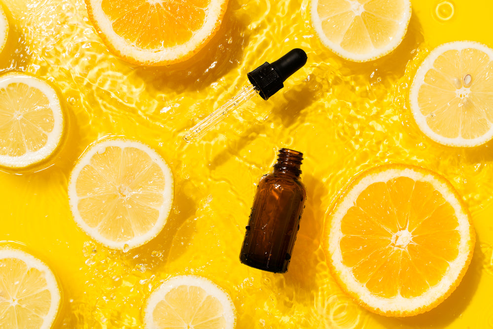 Bottle of Vitamin C serum floating in water with orange and lemon peels against a yellow backrgound