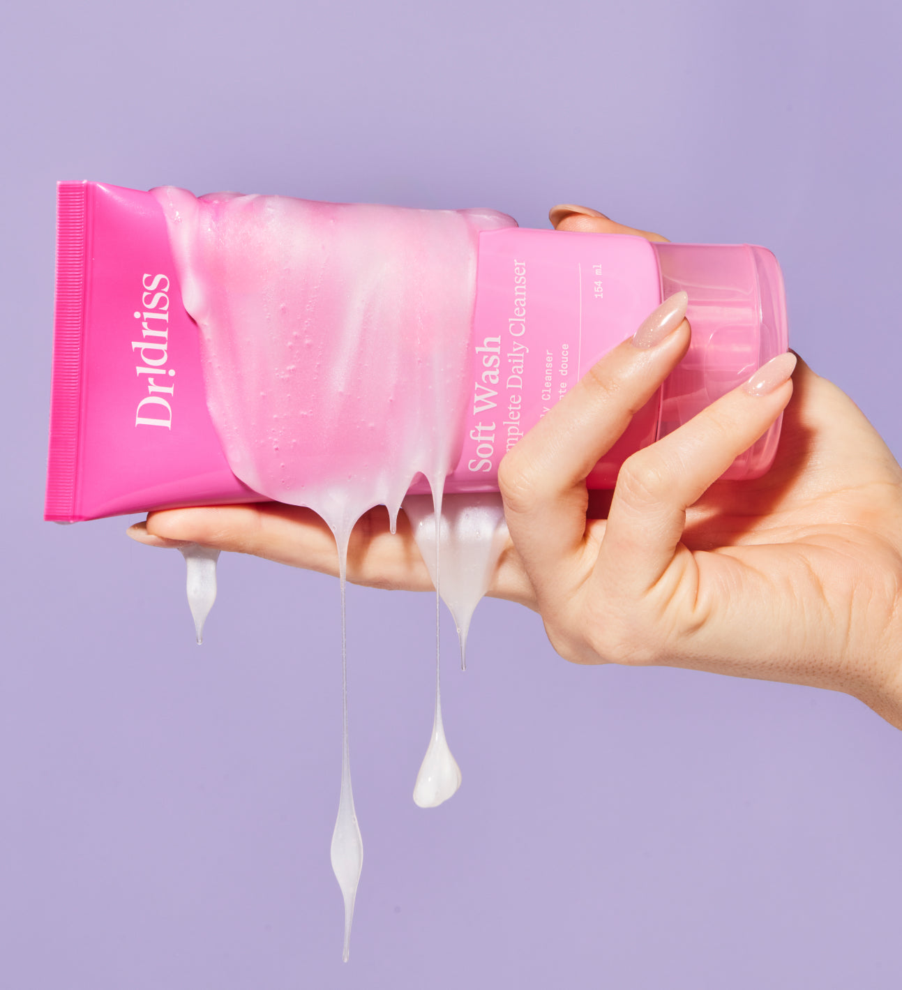 Hand holding a pink package of Dr. Drlriss Soft Wash with white liquid dripping, against a light purple background.