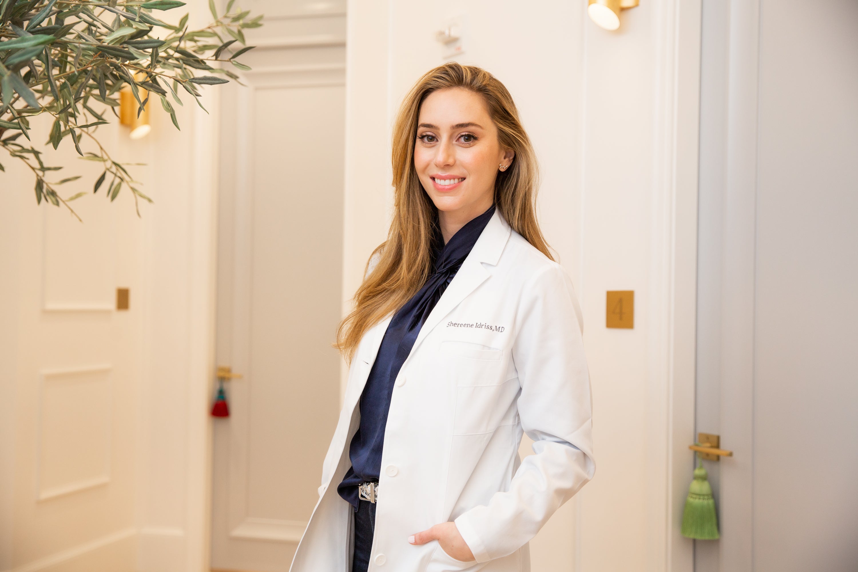 Dermatologist Dr. Shereene Idriss, MD, in a white lab coat and navy blouse, smiling confidently in a modern office setting