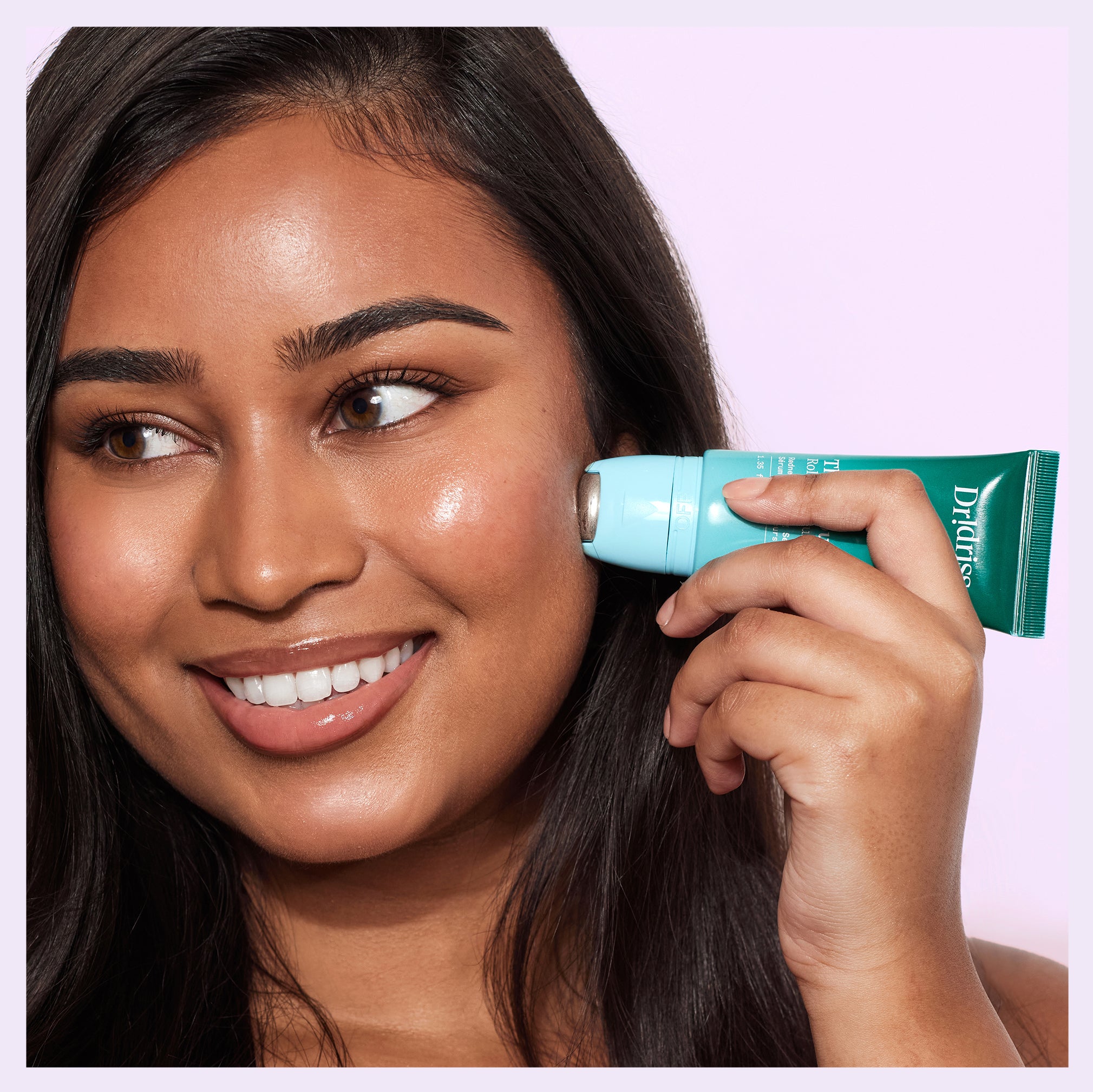 Smiling woman applying Dr. Idriss The Depuffer Roll-On Serum to her cheek, showcasing radiant, calm skin