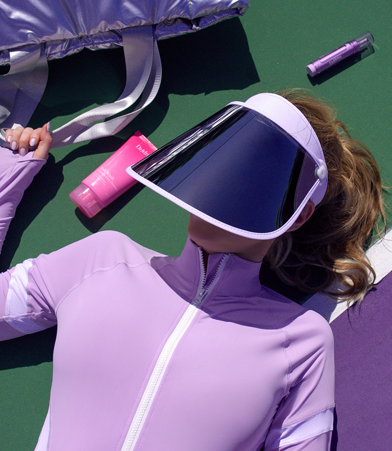 Dr. Shereene Idriss lying on a tennis court wearing the Major Shade Full Lux Sunshield in white, with various Dr. Idriss products lying around her.